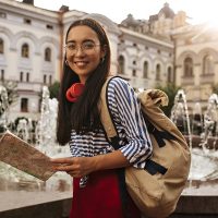 Attractive Pretty Asian brunette woman I silk skirt, striped shirt and eyeglasses looks into camera, smiles. Charming girl holds map and backpack near fountain.