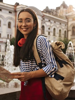 Attractive Pretty Asian brunette woman I silk skirt, striped shirt and eyeglasses looks into camera, smiles. Charming girl holds map and backpack near fountain.