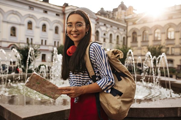 Attractive Pretty Asian brunette woman I silk skirt, striped shirt and eyeglasses looks into camera, smiles. Charming girl holds map and backpack near fountain.